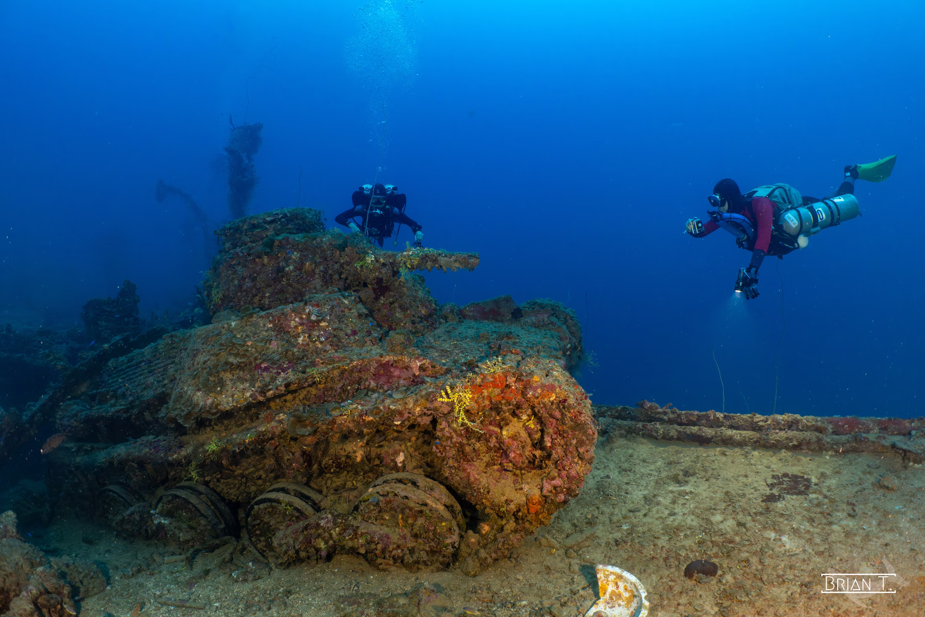 Divers above a coral-encrusted tank resting on a wreck in deep blue water